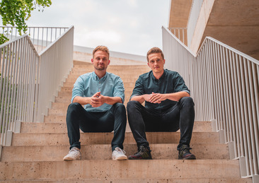 Tobias Düser und Hendrik Sander sitzen auf einer Treppe und blicken in die Kamera